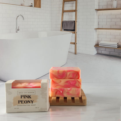 Brick City Scents Pink Peony soap bars stacked on a wood holder with product box in bathroom setting.